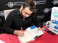 Un hombre en una firma de libros, con un libro abierto en su mano derecha y un bolígrafo en la izquierda. En el fondo, se puede ver una camiseta con el nombre de Jorge Vázquez y un cartel que dice "Fundación Cofares". La mesa es roja y tiene otros libros sobre ella.