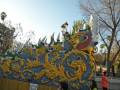 Un carro de carnaval con detalles dorados y azules, una figura de dragón y personajes vestidos de verde. Fondo con árboles, palmeras y un cielo claro.
