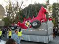Un carro de carnaval con un dragón rojo y blanco en el frente, acompañado por personajes de dibujos animados. La carroza está decorada con detalles de un barco y tiene una gran rueda en la parte frontal. La escena se desarrolla en un ambiente festivo, con personas observando el desfile y árboles al fondo.