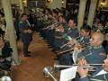 Una banda militar en uniforme toca instrumentos de viento y percusión. El escenario es decorado con columnas y luces, indicando un ambiente formal. La imagen captura la esencia de una ceremonia o concierto militar.
