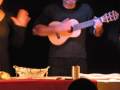 Una pareja de músicos en un escenario, el hombre toca un ukulele mientras la mujer gestiona con una mano. La escena está iluminada por el público que se puede ver en la sombra.