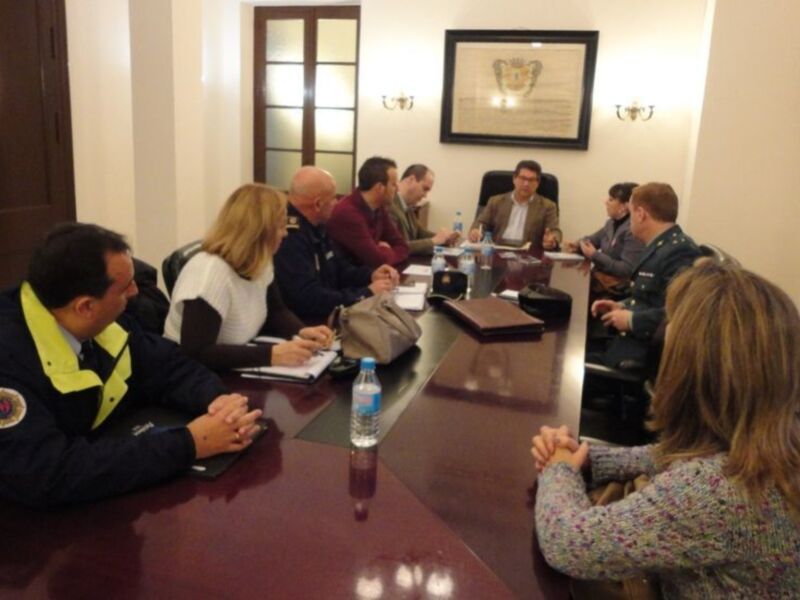 Un grupo de personas en una reunión formal, sentadas alrededor de una mesa grande con documentos y botellas de agua. La sala tiene detalles decorativos en el techo, como lámparas y un marco con una insignia.