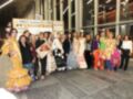 Una fila de modelos posa en un evento de moda, con vestidos de colores brillantes y diseños elegantes. El entorno es moderno con iluminación artificial y una estructura metálica en el fondo.