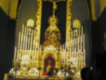 Un altar con una imagen central de la Virgen María, rodeada por candelabros y flores. En el centro de la imagen, hay una cruz con un Cristo crucificado. La iluminación es suave y dorada, creando una atmósfera de reverencia.