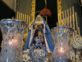 En el centro, una figura vestida con un manto azul se encuentra ante un altar dorado. A su alrededor, hay velas cristalinas y una cruz en el techo de la iglesia. La atmósfera es reverente, con luces suaves que iluminan la escena.