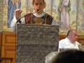 Un sacerdote habla en un altar adornado con mosaicos religiosos. El fondo muestra imágenes de santos y figuras bíblicas, mientras el sacerdote usa un micrófono para dirigirse a una audiencia.
