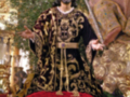 Imagen de un personaje vestido con trajes tradicionales, posiblemente de la época colonial o barroca, en un entorno que parece ser una iglesia. La figura está sosteniendo una vara y tiene un escudo en la espalda, lo que sugiere un papel de liderazgo o autoridad. La escena está iluminada con luces doradas y hay flores dispuestas en el suelo.