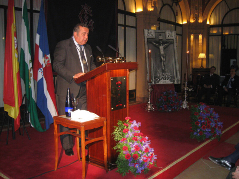 Persona hablando en un evento formal con banderas y flores en el escenario.