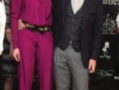 Dos personas posan en un evento formal, uno con traje y corbata, la otra con pantalones largos y una chaqueta roja.