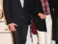 Dos personas posan en un evento, uno con traje y corbata, la otra con blazer y pantalones blancos. El fondo es un cartel de promoción para el "Salón Internacional del Cómic".