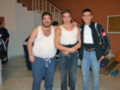 Tres hombres posan juntos en una habitación con escaleras y una puerta visible. El hombre del medio lleva un chaleco de seguridad blanco, mientras que los otros dos están vestidos con camisetas blancas y pantalones de mezclilla. La pared detrás de ellos tiene una pizarra con notas y un cartel.