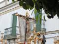 Imagen de una procesión religiosa con figuras en el paso central, un Cristo crucificado y otros personajes vestidos de blanco. El fondo muestra una iglesia con balcones verdes y árboles en primer plano.