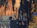 Imagen de una procesión religiosa con un crucificado en el centro, rodeado por velas y decoraciones doradas. Personas vestidas formalmente observan la escena.