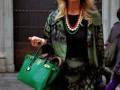 Elegante mujer con abrigo verde y negro, bolso verde y collar multicolor, frente a una puerta de madera tallada.