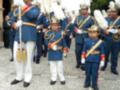 Guardias formales en uniforme azul y blanco, con insignias doradas. Niños pequeños también vestidos en uniforme similar, posiblemente como parte de una ceremonia o desfile. Escenario al aire libre con vegetación y estructuras en el fondo.