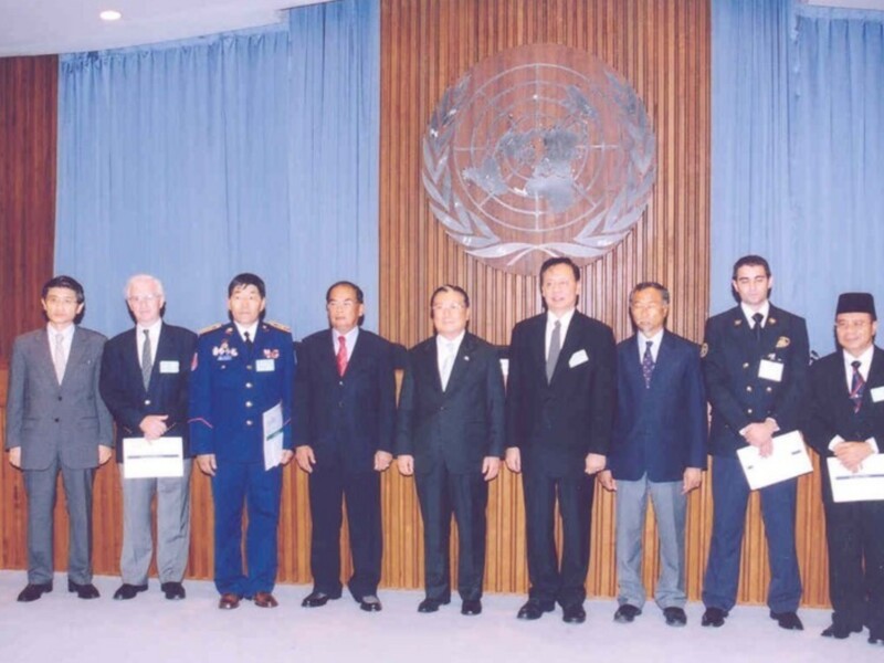 Una imagen de una ceremonia oficial con varios hombres vestidos formalmente, posando frente a un escudo de armas que representa la ONU. Algunos hombres llevan insignias militares, lo cual sugiere que pueden ser oficiales de la fuerza armada. La escena está ambientada en un entorno formal con cortinas azules y una pared de madera.