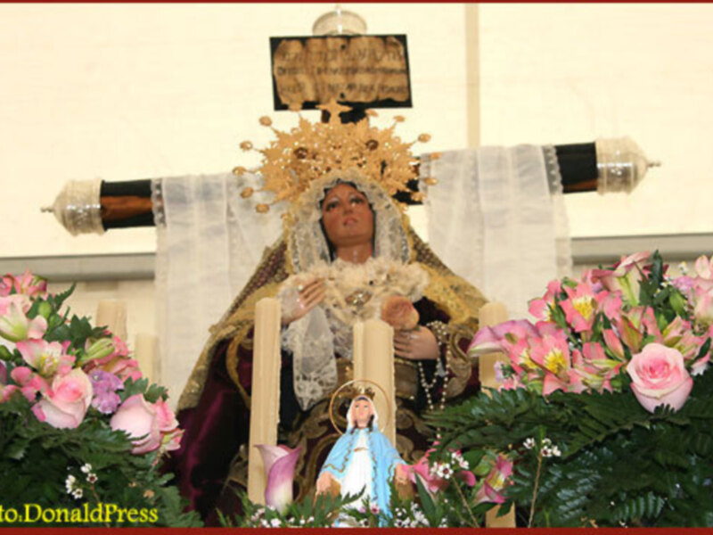 Imagen de la Virgen María adornada con flores y joyas, rodeada de ofrendas florales y decoración festiva.