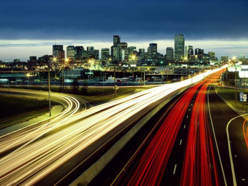 Carretera iluminada con trazos de luces de coches en movimiento, con edificios urbanos iluminados al fondo al atardecer.