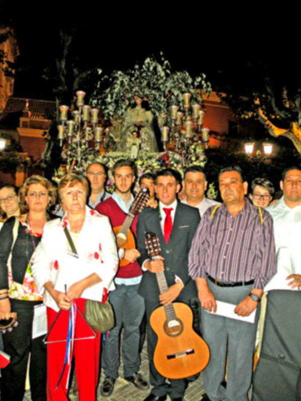 Una imagen festiva nocturna con una multitud de personas vestidas formalmente, algunos con sombreros y otros con instrumentos musicales. La escena está iluminada por faroles, enfocándose en un altar o estatua central rodeado de flores y velas. La celebración parece ser una festividad religiosa o cultural importante.