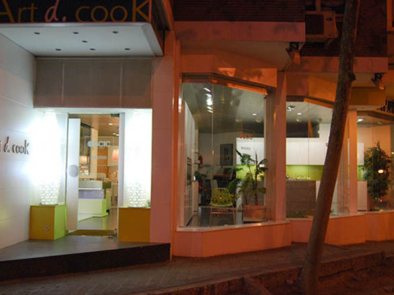 Exterior de la tienda 'Art & Cook' iluminada por la noche, mostrando su entrada y vitrinas con plantas y muebles modernos.