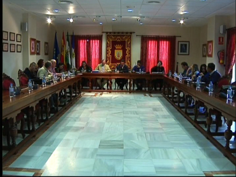 Sala de reuniones con mesa central y personas sentadas en sillas alrededor, decorada con banderas y cortinas rojas.