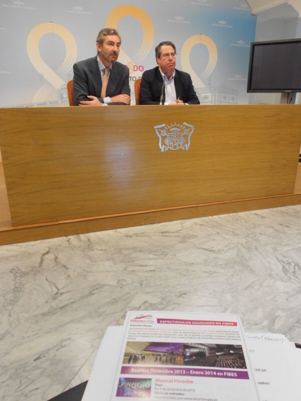 Dos hombres en traje sentados detrás de un mostrador con el escudo de Sevilla, promocionando espectáculos navideños en FIBES.
