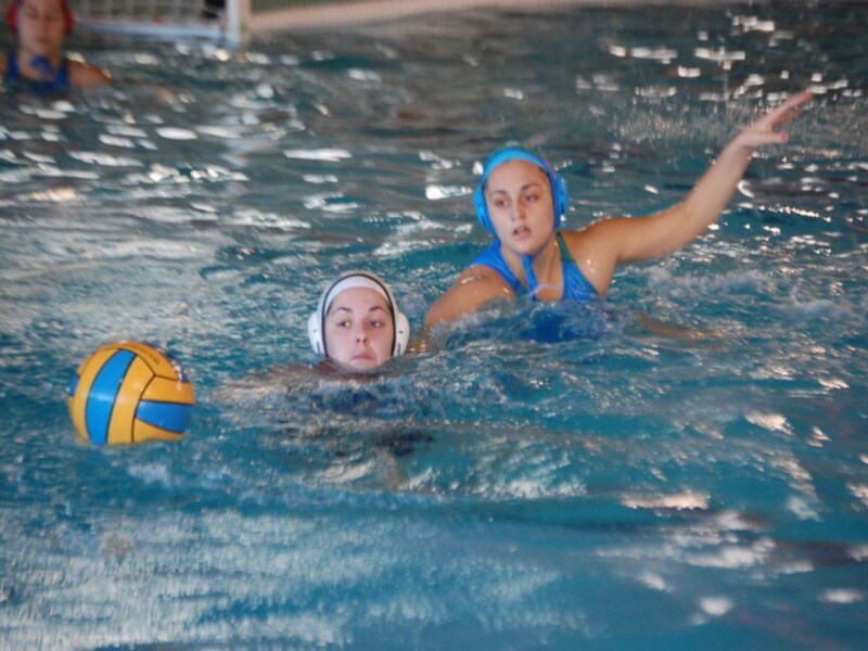 Jugadoras de waterpolo en acción, una lanza el balón mientras la otra la persigue en una piscina.