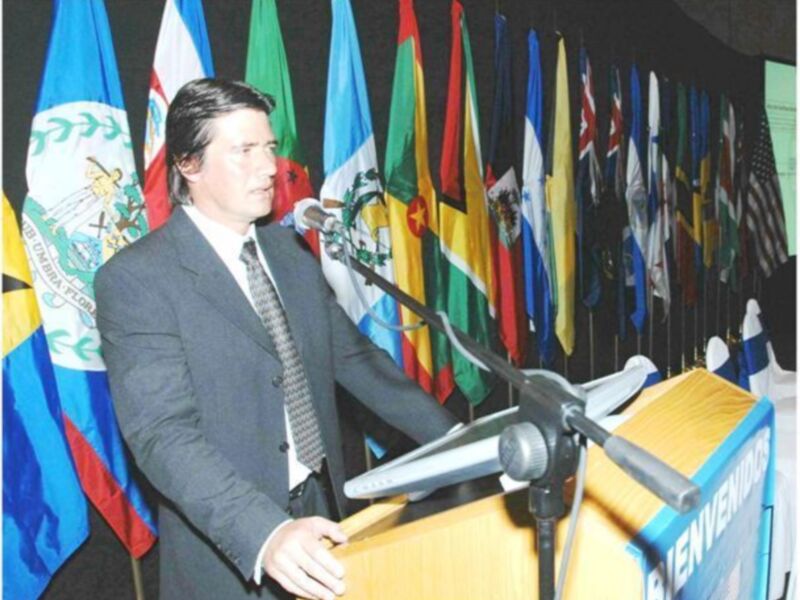 Persona hablando en un evento formal con banderas de varios países en el fondo.