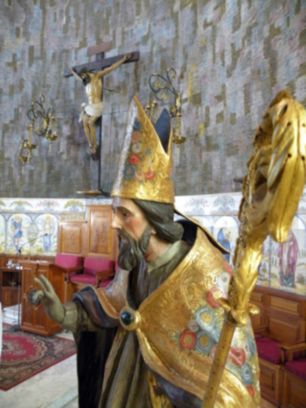 Figura religiosa dorada con manto y cetro, en un interior con decoración barroca y crucifijo en el fondo.