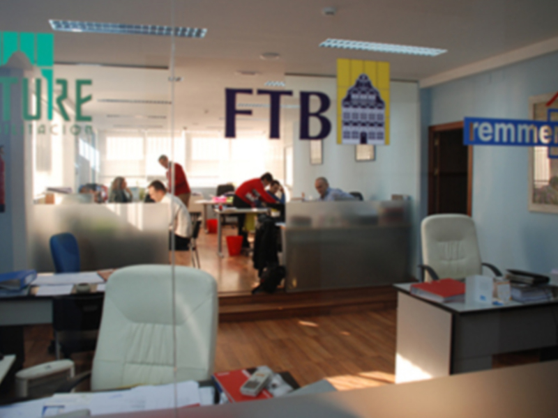 Oficina FTB con empleados trabajando en sus mesas, logo y muebles de oficina visibles.