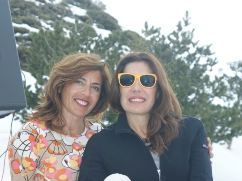 Dos mujeres sonríen en un paisaje nevado con árboles de pino en el fondo.