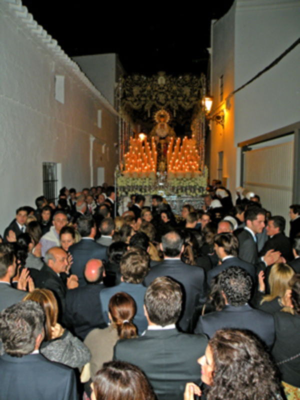 Celebración religiosa nocturna con una imagen procesional adornada y una multitud congregada en una calle iluminada.