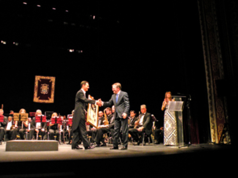 Dos hombres se saludan en una escena formal, posiblemente en un evento de premiación o ceremonia, con un fondo oscuro y una silla en el escenario.