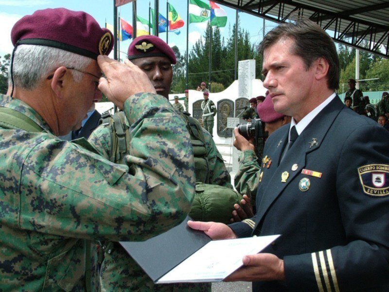 Entrega de reconocimiento en ceremonia militar con banderas internacionales en el fondo.
