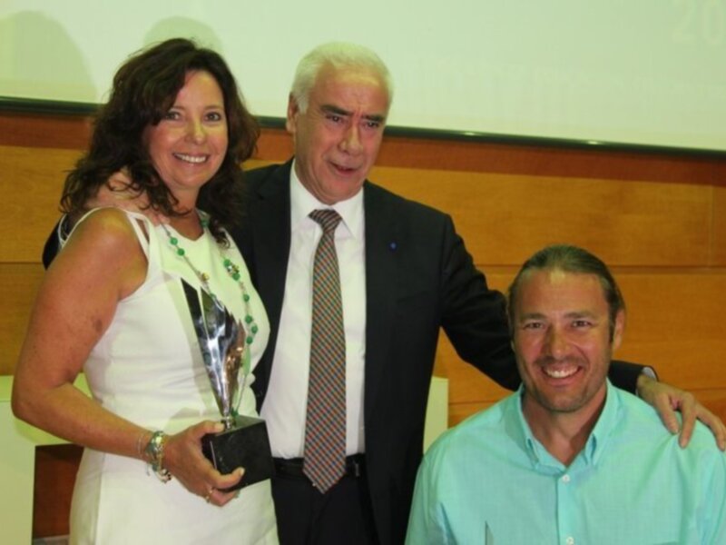 Persona recibiendo un trofeo en un evento formal, acompañada por dos individuos en un escenario con proyección de '2013' en el fondo.
