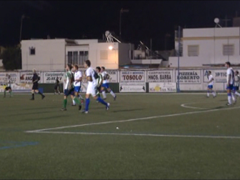 Un partido de fútbol nocturno en un campo iluminado con jugadores en acción. La imagen muestra a varios jugadores corriendo y luchando por el balón en un campo de juego con líneas blancas. La iluminación del estadio brilla sobre el césped verde, y en el fondo se pueden ver edificios y anuncios publicitarios.