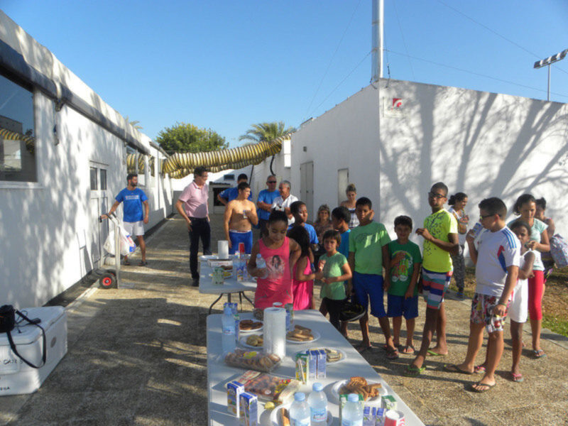 Un grupo de personas, principalmente niños y adultos, se reúnen alrededor de una mesa con comidas y bebidas. La escena está en un área abierta, frente a una estructura blanca con una palmera y un poste de luz en el fondo. La gente parece estar disfrutando de una reunión o evento al aire libre, con la mesa llena de alimentos y bebidas.