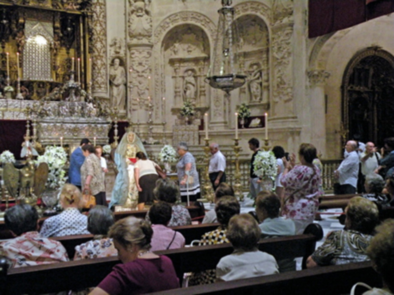 Interior de una iglesia con altares ornamentados, estatuas religiosas y flores. Personas en vestimenta tradicional observan el altar central, mientras otros se sienten en bancos. La arquitectura barroca destaca con detalles dorados y marmolados.