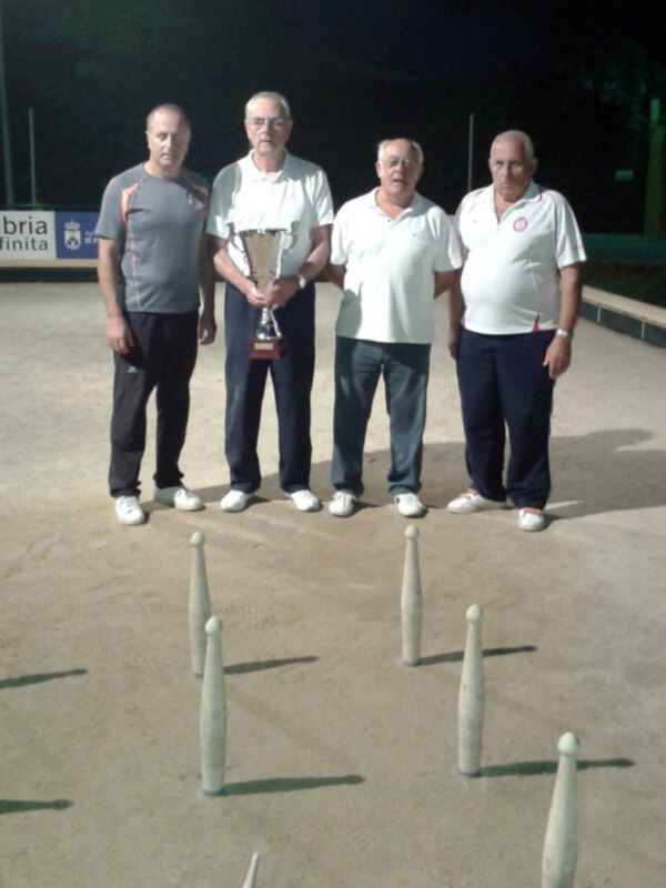 Un grupo de personas posando con un trofeo, posiblemente en un torneo de boliche, con una pista de boliche en el fondo.