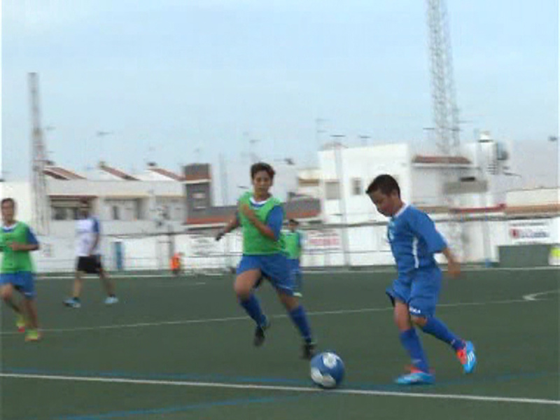 Jóvenes jugando fútbol en un campo deportivo al aire libre, con edificios y torres de fondo.