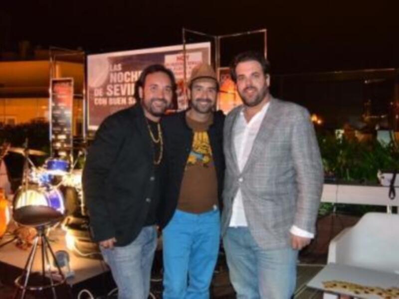 Tres hombres sonrientes posando juntos en un evento nocturno con música y decoración de luces.