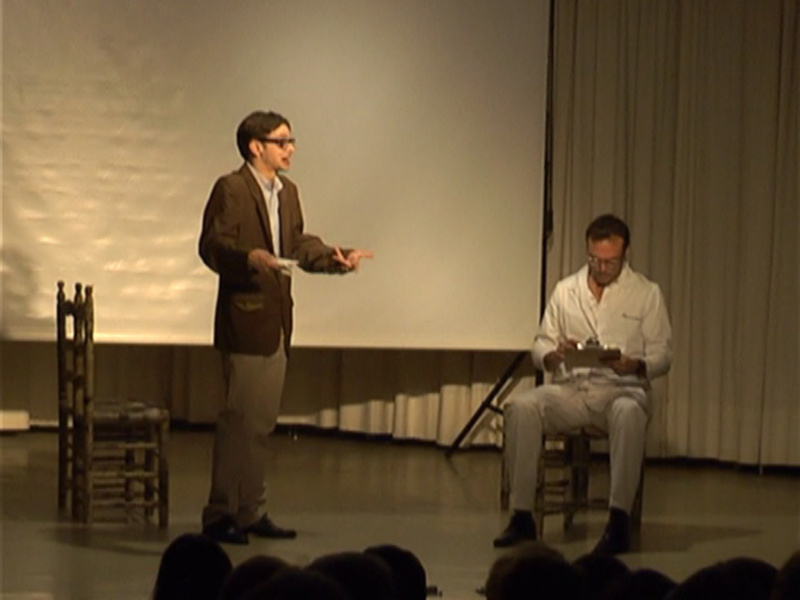 Un hombre en una presentación en un escenario, mientras otro hombre se sienta en una silla, parece estar leyendo o escribiendo algo en un bloc de notas.