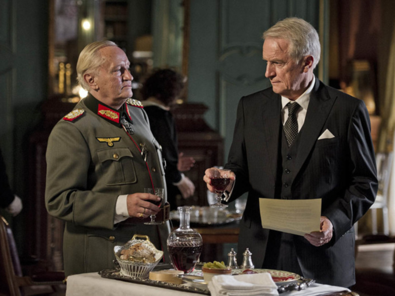 Dos hombres en trajes de gala, uno con uniforme militar, en un ambiente formal con copas de vino y pasteles en la mesa. La escena sugiere una reunión de alto nivel o una celebración formal.