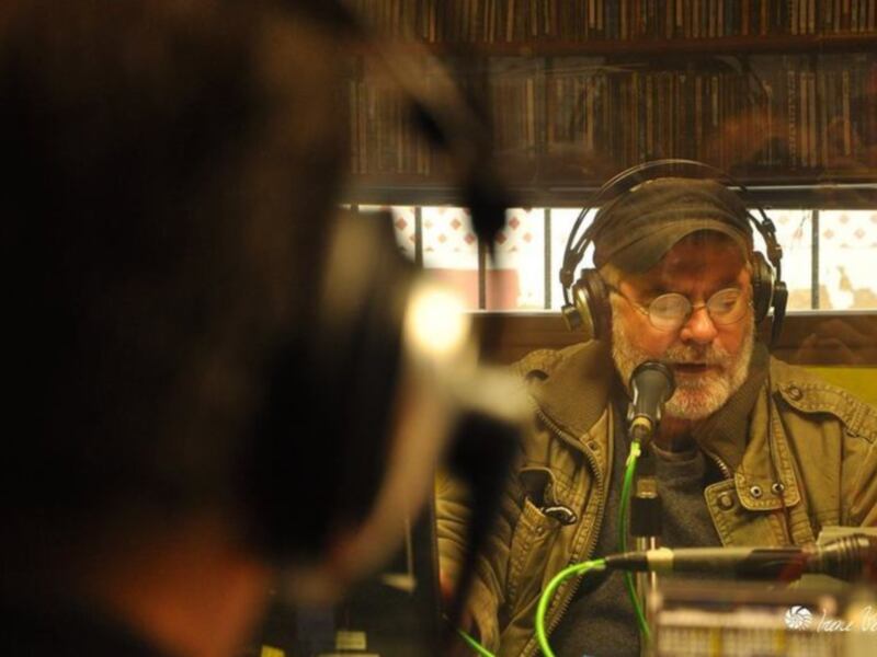Un hombre con auriculares y una chaqueta verde está en un estudio de radio, hablando en un micrófono. Al fondo, se pueden ver estanterías con libros y una ventana con cortinas de lunares.