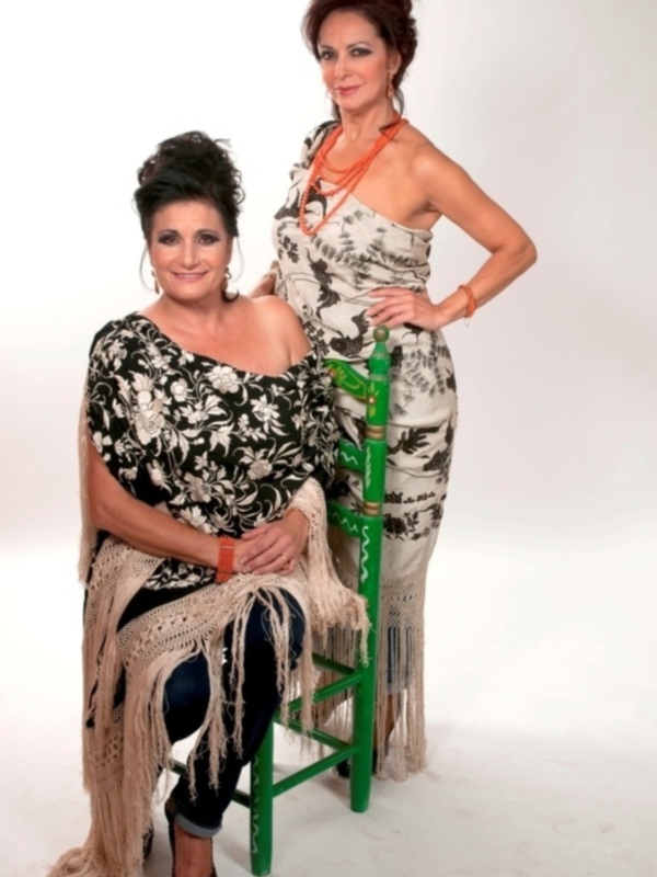 Dos mujeres posando con elegancia y estilo en un estudio fotográfico, una sentada y la otra de pie, ambas con vestidos y accesorios llamativos.