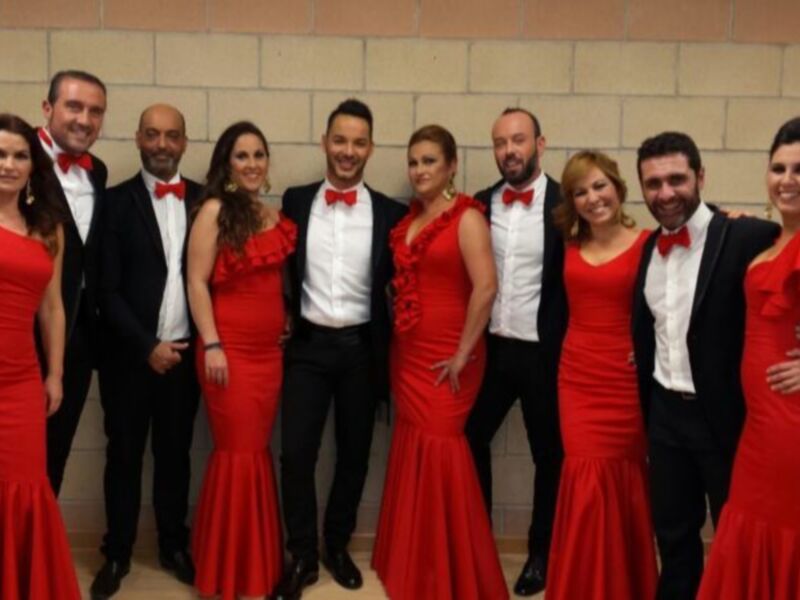 Una imagen de una banda de música en un escenario, con los músicos vestidos formalmente y las mujeres con vestidos rojos.