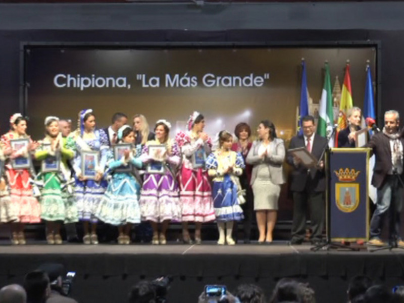 Una celebración en Chipiona con personas en trajes tradicionales y una pantalla que dice 'Chipiona, La Más Grande' en el escenario.
