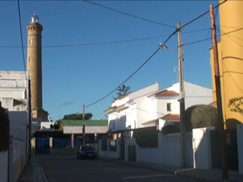 Imaginación en la calle, con una torre de madera que se eleva hacia el cielo azul. La imagen muestra una calle con edificios de diferentes colores, incluyendo un edificio amarillo y blanco. También se puede ver una línea de cables eléctricos que cruzan el cielo. La escena es tranquila, con un ambiente soleado y una vista clara de la torre.