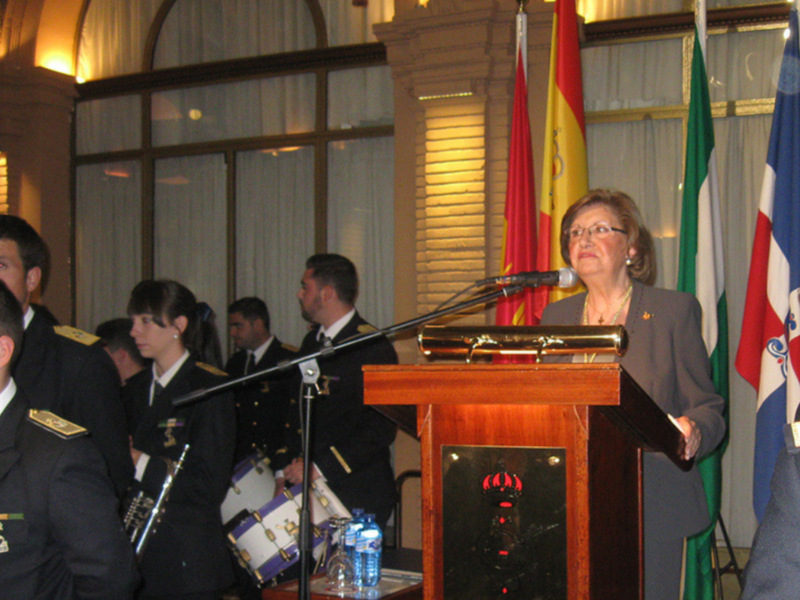 Mujer hablando en un podium en una ceremonia formal con banderas de varios países y uniformes militares al fondo.