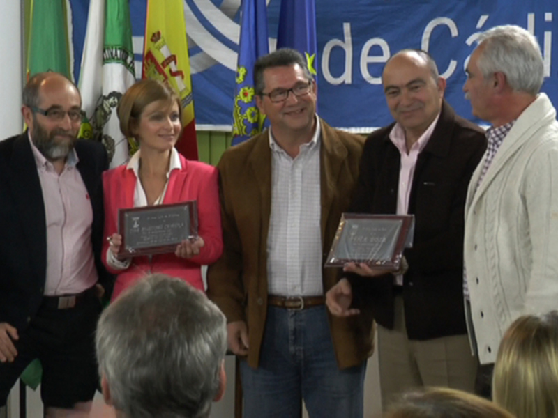 Una celebración formal donde una mujer sostiene un reconocimiento junto a otros individuos en una sala con banderas y un banner en el fondo.
